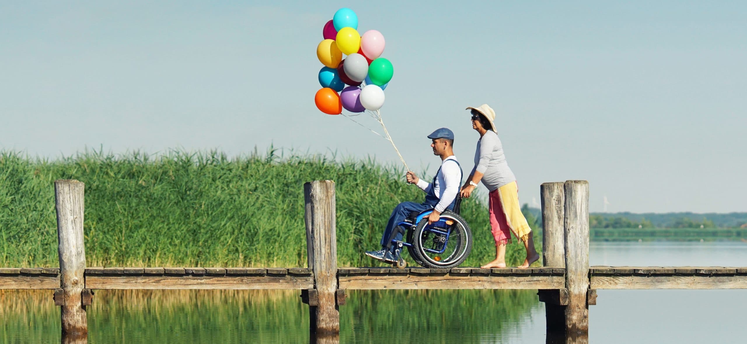 Frau schiebt einen Mann im Rollstuhl mit bunten Luftballons über einen Holzsteg am Wasser.