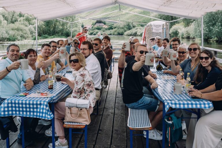 Teamfoto der sowis GmbH beim Sommerfest 2025 auf einem Holzfloß vor einem See mit blau-weiß karierten Tischen, lachend und anstoßend mit Bierkrügen.