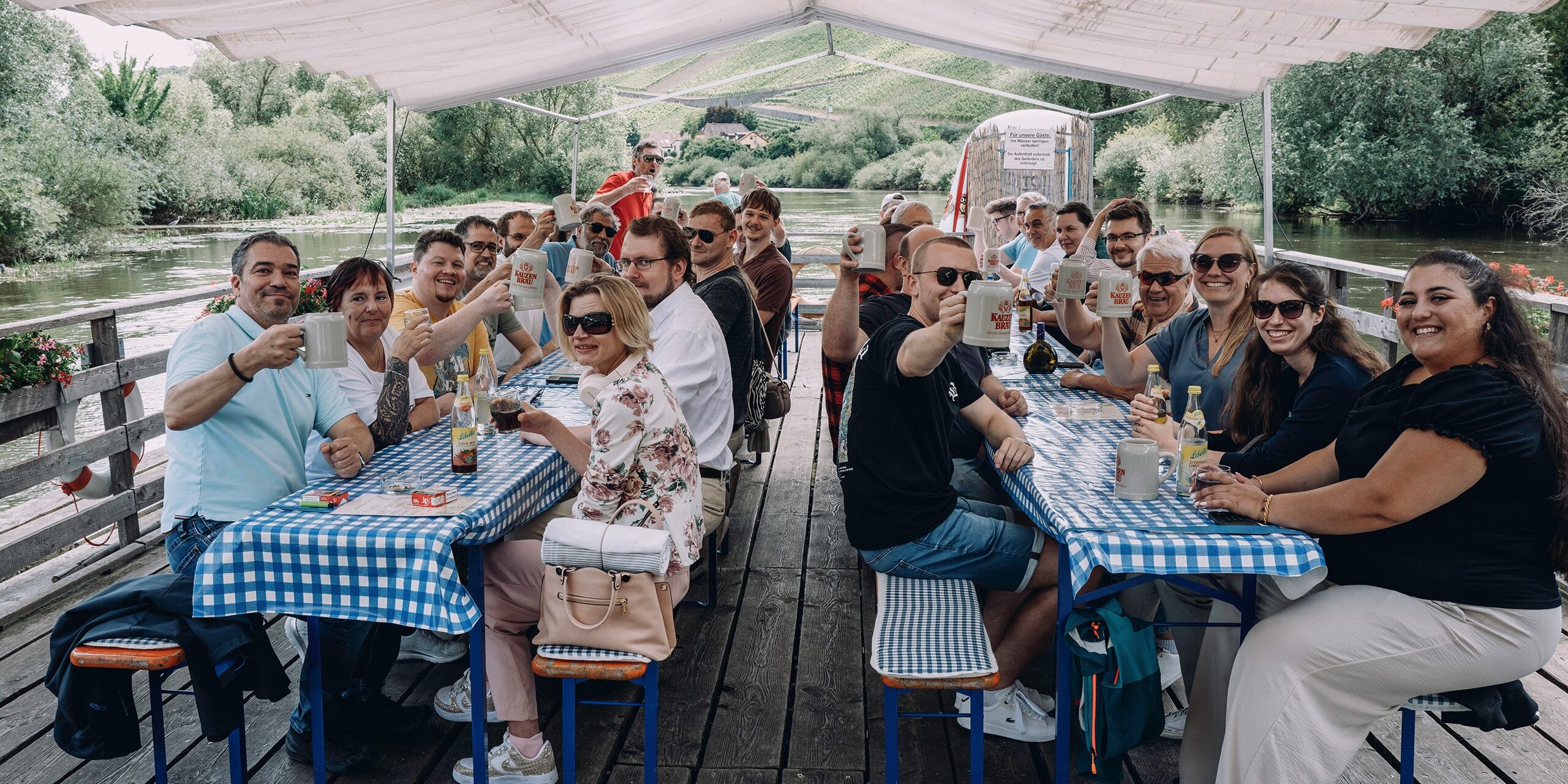 Teamfoto der sowis GmbH beim Sommerfest 2025 auf einem Holzfloß vor einem See mit blau-weiß karierten Tischen, lachend und anstoßend mit Bierkrügen.